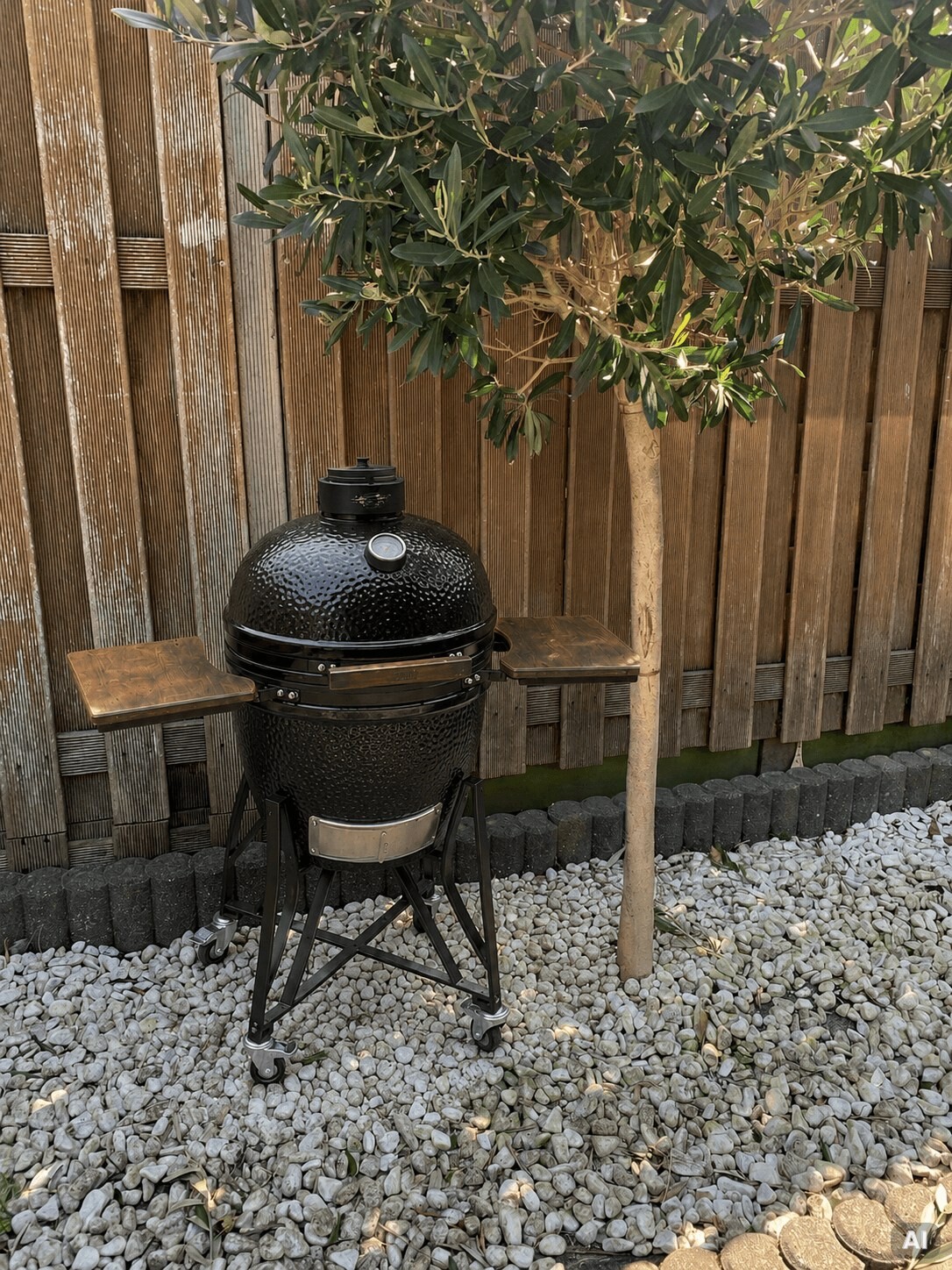 Kamado Grill zwischen Holzzaun und Olivenbaum auf der Terrasse.